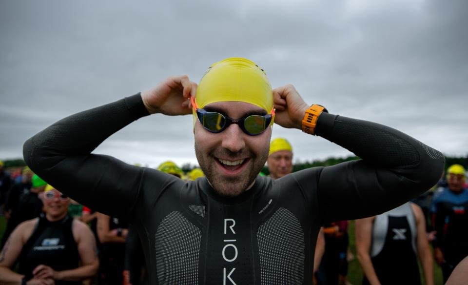 A smiling triathlete in a black wetsuit adjusts his goggles and yellow swim cap while preparing for the start of the race.