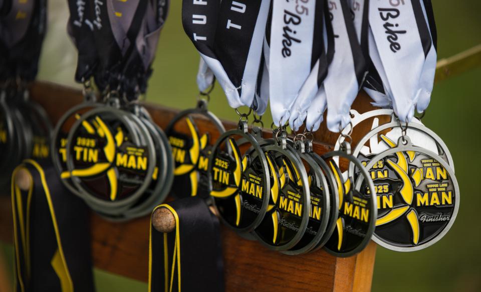 A row of silver 2025 Tupper Lake Tinman finisher medals with yellow and black accents hang from ribbons on a wooden rack.