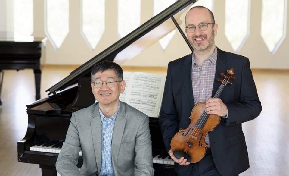 A man sitting on the bench in front of a grand piano with a man standing next to him holfing a violin