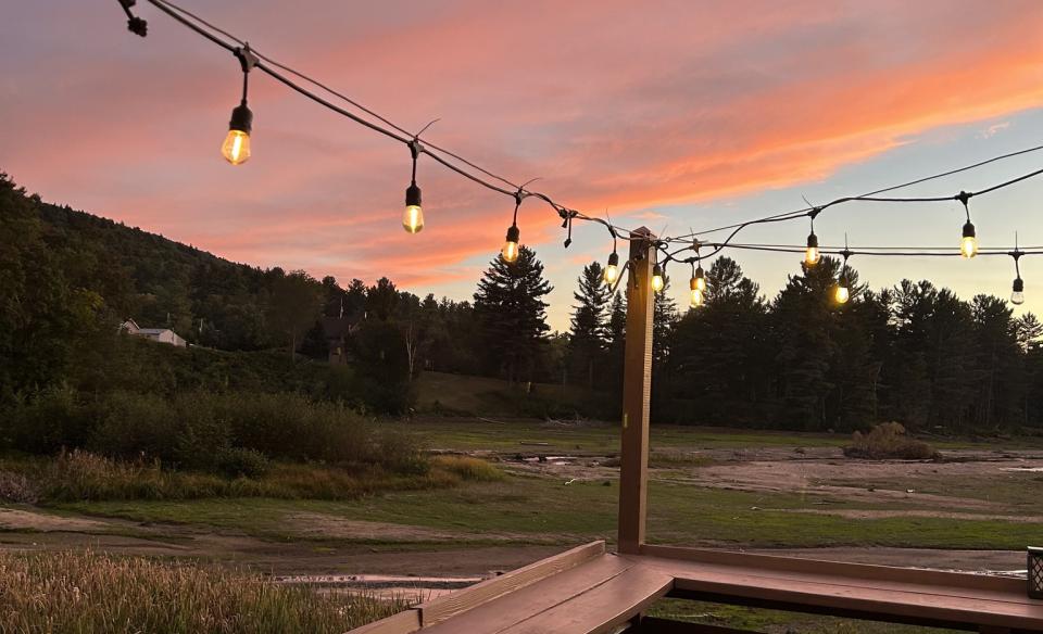 view from a wooden deck with party lights strung above looking out to trees and a sunset with orange and pink colored clouds
