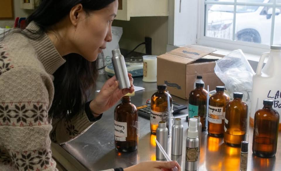 A woman smells a small silver bottle while surrounded by various amber-colored bottles and other containers of liquid on a stainless steel counter.