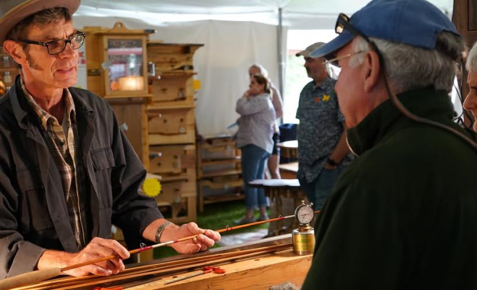 A man in a dark jacket and hat shows a handcrafted fishing rod to two people.