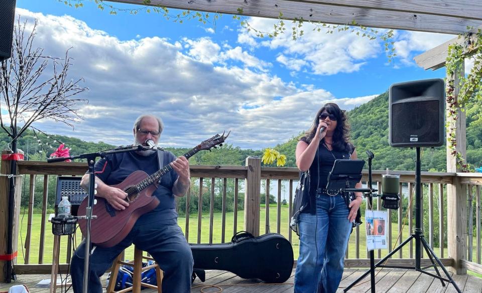 A man and woman perform music on an outdoor deck. The man plays an acoustic guitar and the woman sings into a microphone.  They are positioned in front of a scenic view and audio equipment.
