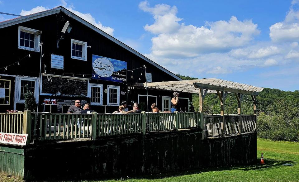 A black building with a large deck and people sitting at tables.  The building has a sign that says The Oxbow Inn.  There is a grassy area in front of the building.