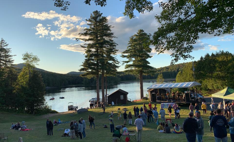Outdoor concert by a lake with people sitting and standing on the grass listening to a band on a stage.