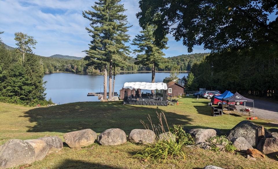 Outdoor stage set up on a lakeside lawn with a few cars parked nearby.