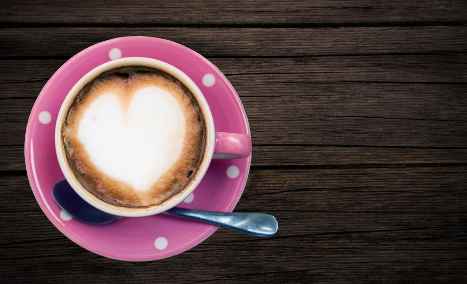 A cappuccino with heart-shaped foam sits in a pink polka-dot cup and saucer on a wooden table
