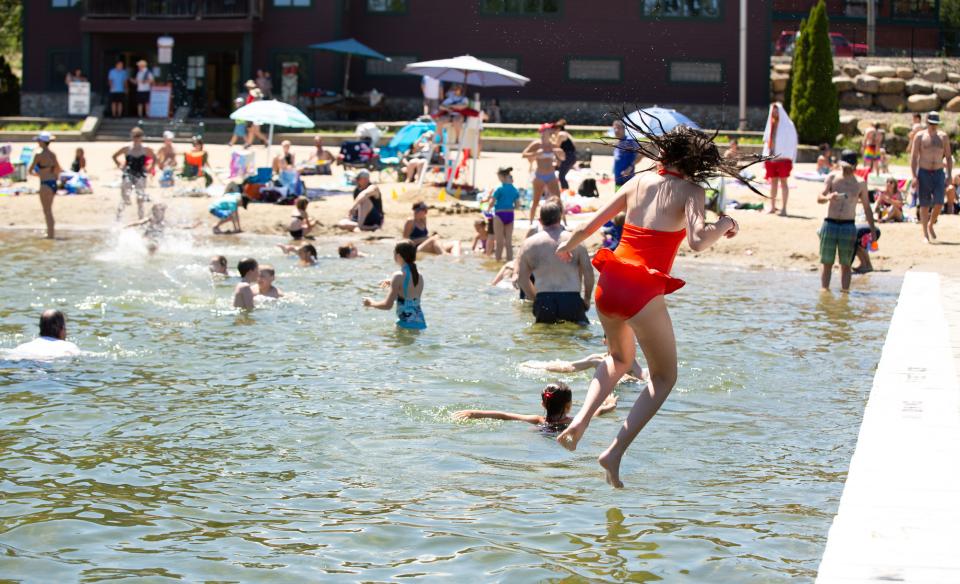 A girl in a red swimsuit jumps into a lake at a crowded beach.  Many people are swimming