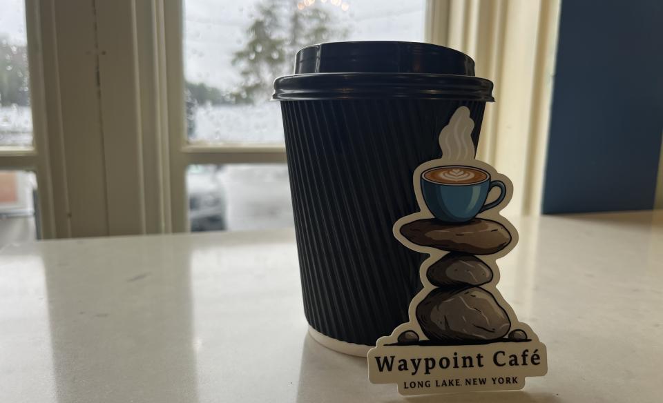 A hot cup of coffee on a table in a to-go cup with custom Waypoint Cafe sticker leaning against it.