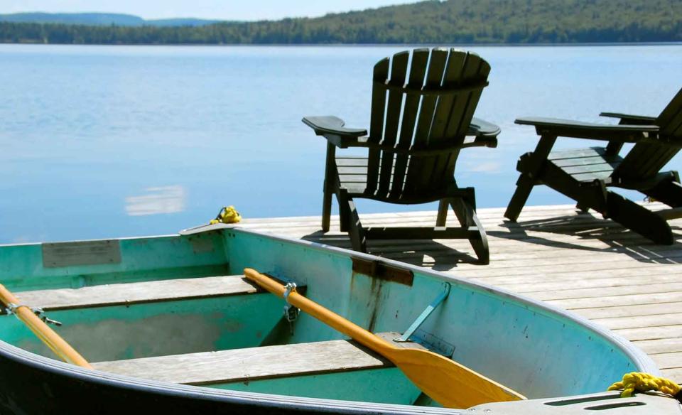 Adirondack chairs on the end of a dock over look the glass like surface of the lake as a blue paddle boat floats nearby