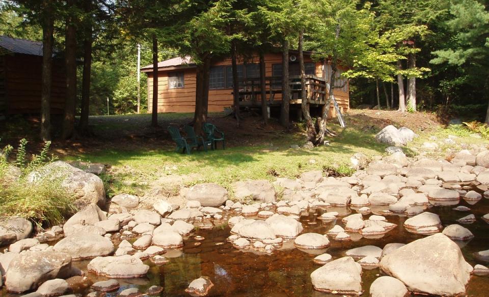 An image of a quaint cottage surrounded with trees and a rocky rivers edge.