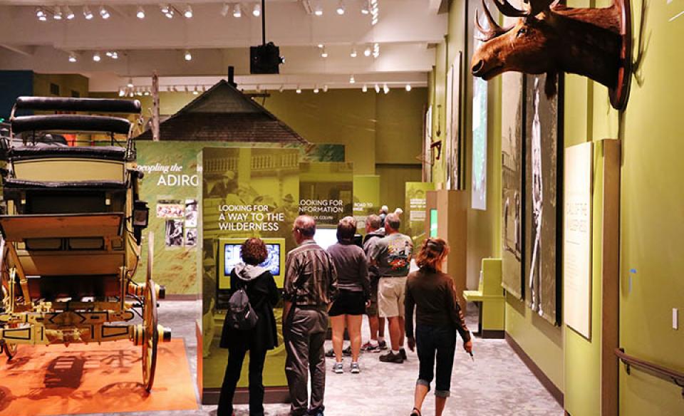 A large room with green walls and high ceilings where people are viewing the plaques under various displays like a carriage and a moose.