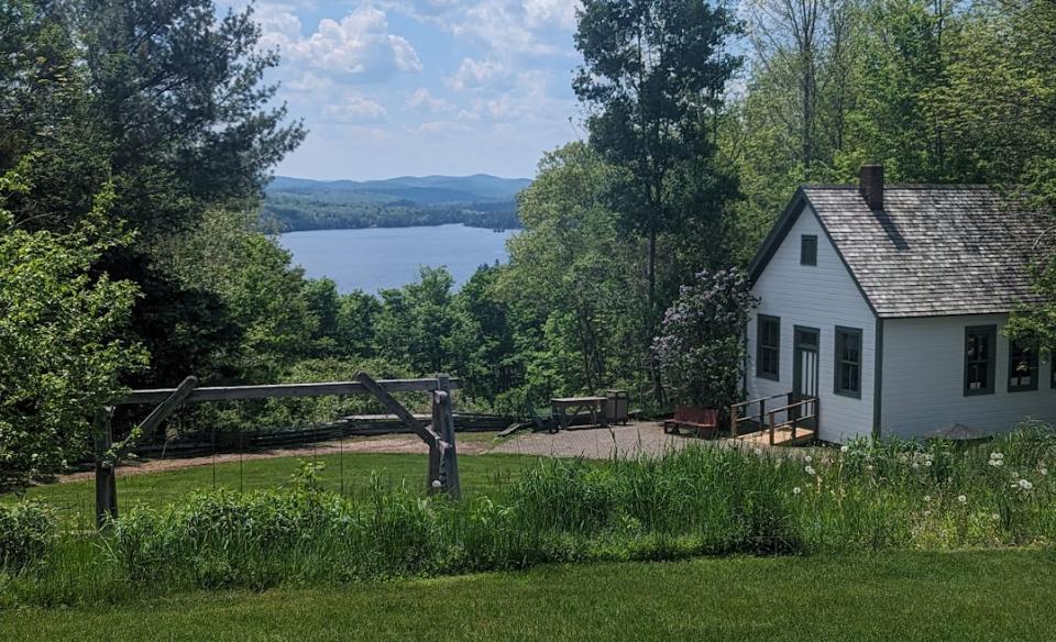 Blue Mountain Lake under billowing white clouds across a lush green lawn in front of a quaint white cabin.