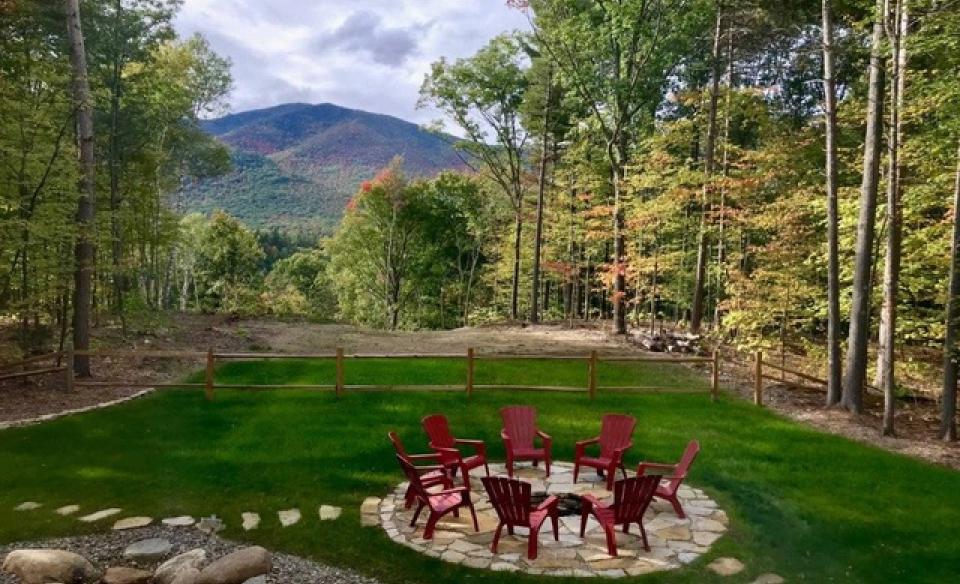 A lush and expansive green lawn with red adirondack style chairs arranged on a patio with fire pit and views of the mountains beyond the trees.