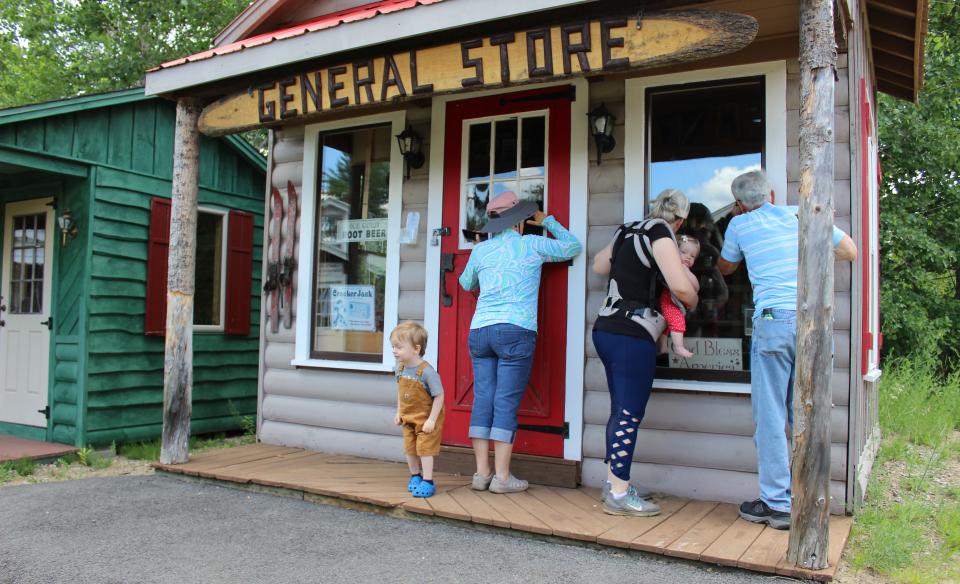 Adults and small children on the porch of a miniature wooden general store.